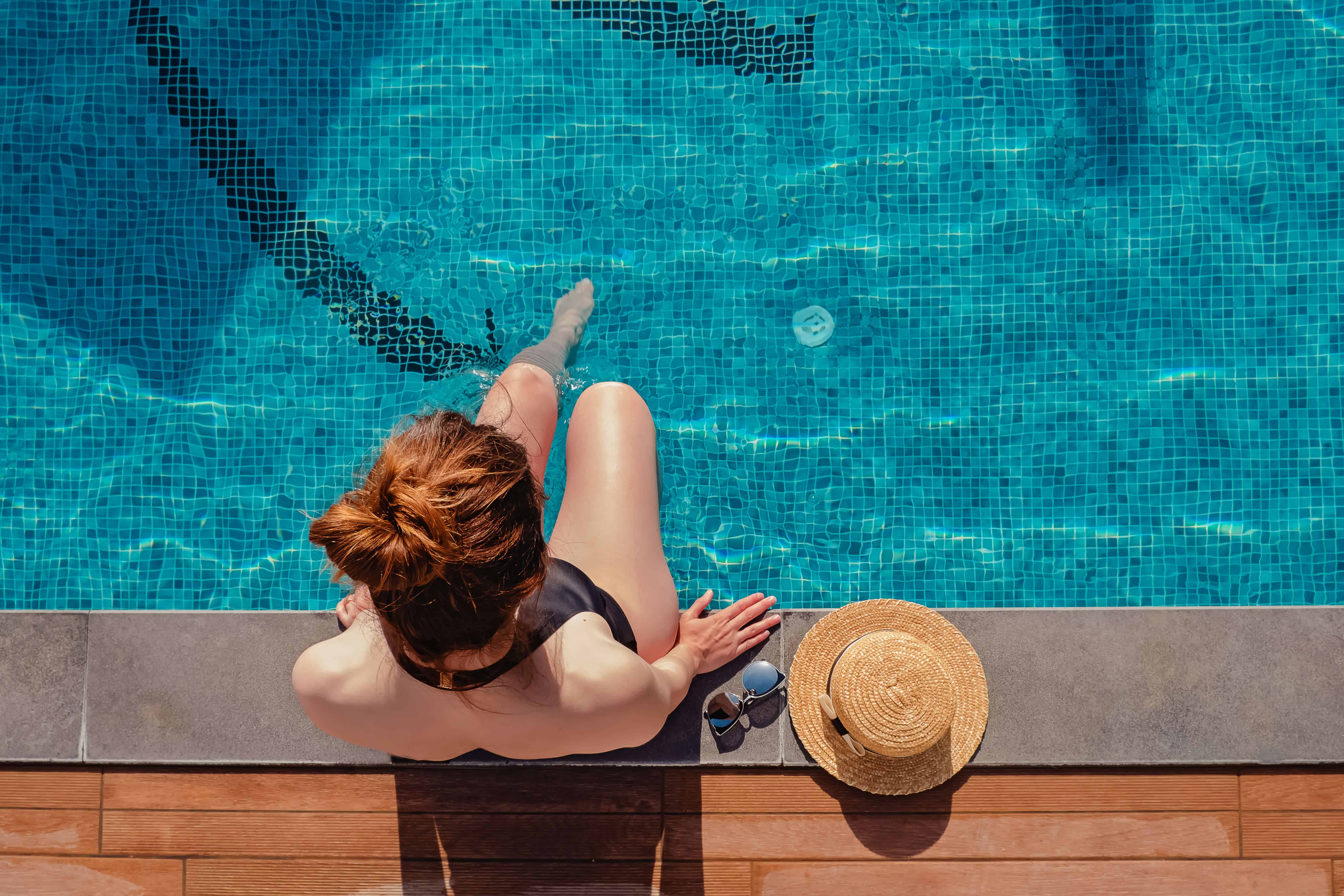 young-woman-sitting-edge-swimming-pool-with-her-straw-hat-lying-near-top-view 2